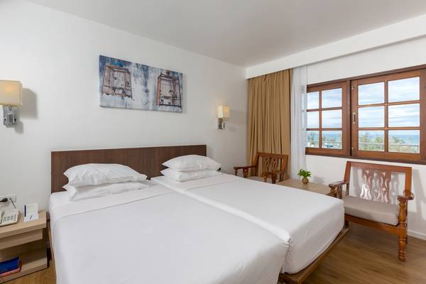 Zwei Betten im Hotelzimmer mit Meerblick, weiße Bettwäsche und Kissen, neben einem Nachttisch und einem Fenster mit Meersicht.