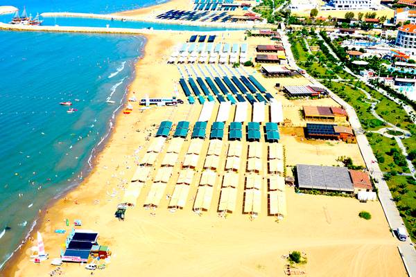 Luftaufnahme des Port Side Resorts, das an einem Strand liegt, mit vielen Sonnenschirmen und Sonnenliegen.