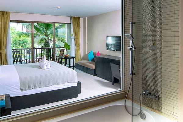 Ein Schlafzimmer mit Badezimmer im Apsara Beachfront Resort und Villa, das eine große Badewanne, ein Bett und einen Balkon mit Aussicht auf einen Baumgarten zeigt.