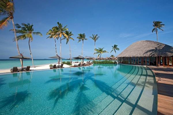 Constance Halaveli Maldives Pool mit Strand, Palmen und einer Strandhütte im Hintergrund.