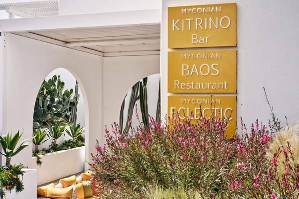 Außenbereich des Myconian Korali Relais & Chateaux Hotels mit Pflanzen und gelben Schildern für Kitrino Bar, Baos Restaurant und Eclectic.