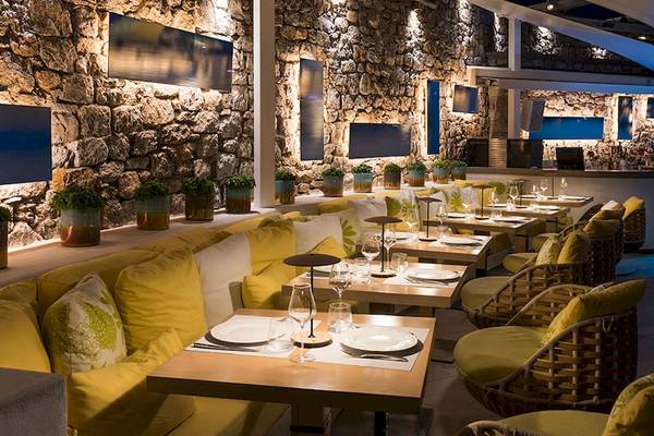 Innenbereich des Restaurants des Myconian Korali Relais & Chateaux mit Steinwänden, gelben Kissen und blauer Beleuchtung.