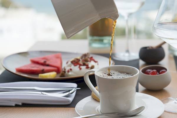 Frühstückstisch im Hotel Myconian Korali Relais & Chateaux mit Kaffee, Wasserglas und Teller mit Wassermelonen, Joghurt und anderen Speisen.