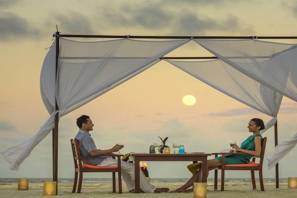 Ein Paar sitzt unter einem Baldachin am Strand und genießt ein romantisches Abendessen bei Sonnenuntergang im Angsana Velavaru.