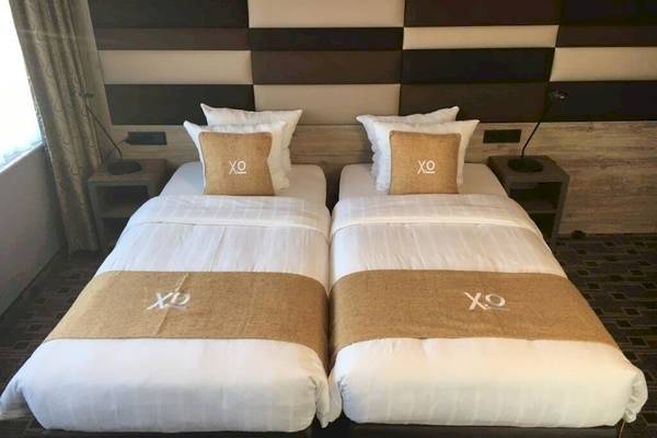 Zwei Doppelbetten im XO Hotels Blue Square Zimmer mit weißen Laken, braunen Kissen und XO-Logo.