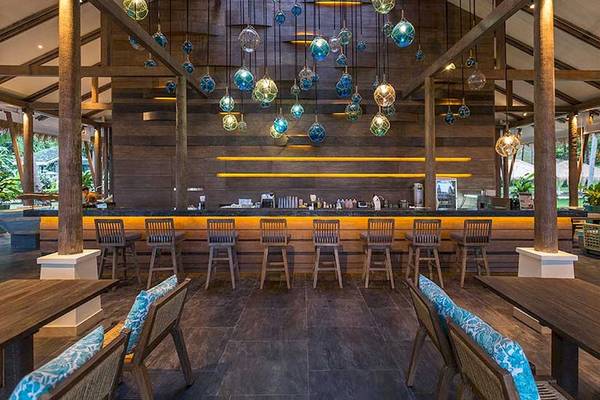 Innenansicht der Bar im Cape Panwa Hotel mit Holzmöbeln und blauen Glasleuchten.
