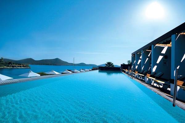 Außenpool des Elounda Bay Palace Hotels am Strand mit weißen Sonnenschirmen und Blick auf das Meer.