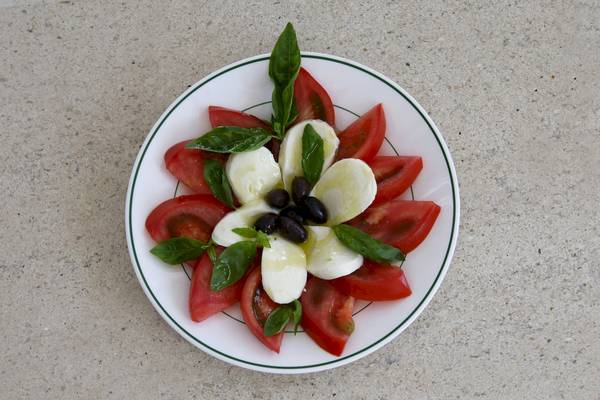 Ein weißer Teller mit Tomatenscheiben, Mozzarella und Oliven, alles in einem Blumenmuster angeordnet.