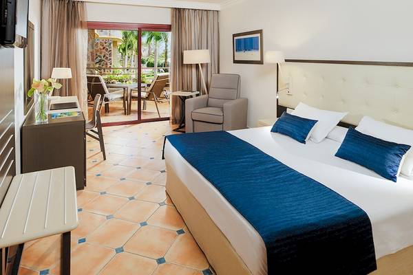 Hotelzimmer im H10 Playa Meloneras Palace mit einem Doppelbett, einem Schreibtisch, einem Sessel und einer Terrassentür.