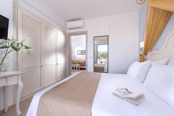 Innenansicht eines Schlafzimmers im Alexander Beach Hotel & Village Resort mit einem Bett, Büchern und einem Tisch mit Blumen.