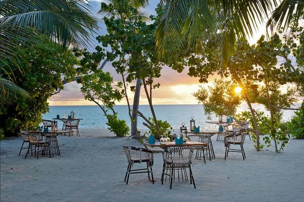 Außenbereich des Kandima Maldives Hotels mit Tischen und Stühlen unter Bäumen am Strand bei Sonnenuntergang.