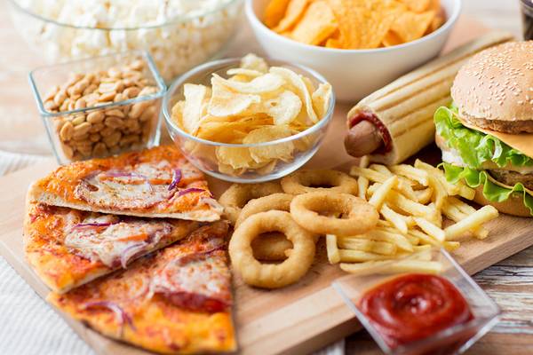 Eine Auswahl an ungesundem Essen wie Pommes, Pizza, Burger, Chips und Soda auf einem Holztisch.