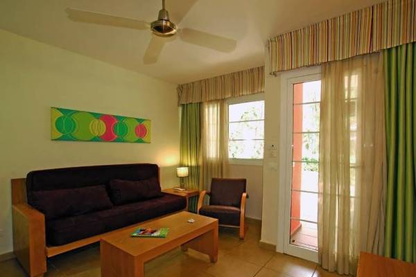 Wohnbereich im Bungalows Cordial Green Golf Hotel mit Sofa, Tisch, Lampe und Fenstern.