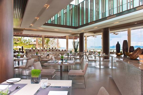 Das Innere des Westin Siray Bay Resort & Spa, Phuket mit vielen Tischen und Stühlen, großen Fenstern und Ausblick auf das Meer.