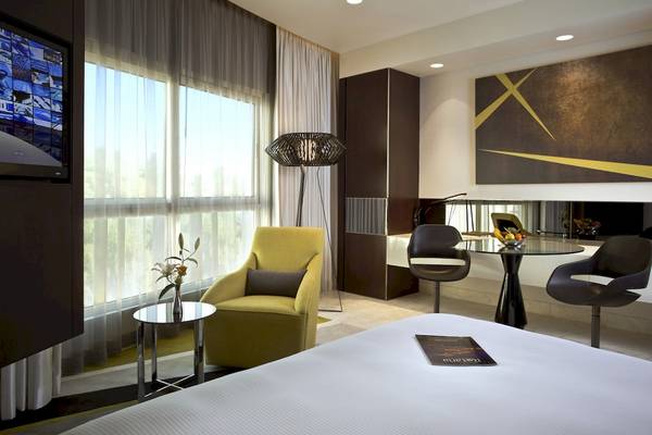 Innenansicht eines Zimmers im Al Ain Rotana Hotel mit Fernseher, gelbem Sessel, Tisch und Kunstwerk an der Wand.