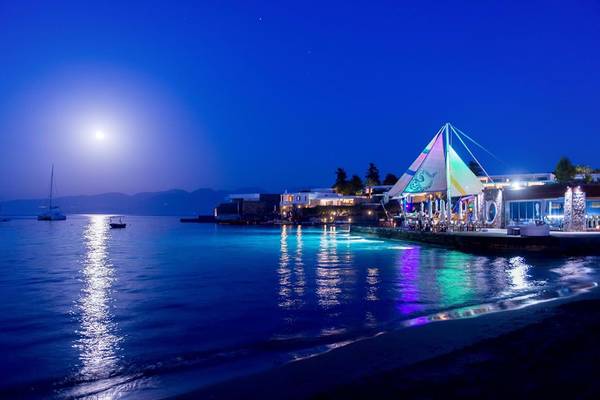 Elounda Beach Hotel & Villas bei Nacht mit Vollmond und farbigen Lichtern am Wasser.