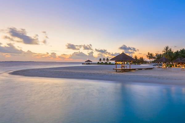 Der Strand des Velassaru Maldives bei Sonnenuntergang mit leuchtenden Lichtern und Pavillons.