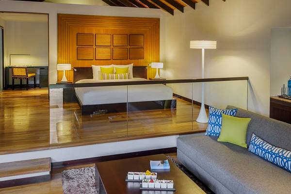 Innenansicht einer Suite im Velassaru Maldives Hotel mit Doppelbett, Wohnzimmerbereich und Glasboden.