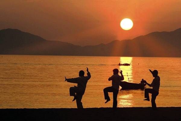 Drei Personen üben Tai Chi auf dem Strand bei Sonnenuntergang.