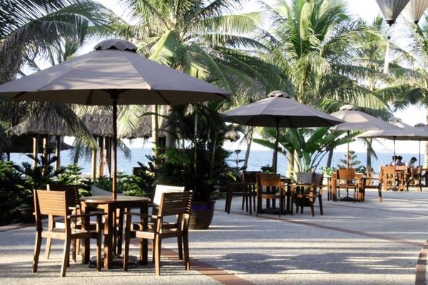 Terrasse des Palm Garden Beach Resort Hoi An mit Tischen und Stühlen unter Schirmen vor einer Palmenkulisse.