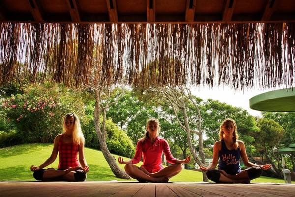 Drei Frauen praktizieren Yoga im Freien auf einer Holzterrasse unter einem Strohdach.