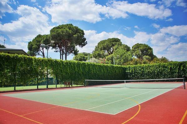 Tennisplatz im Freien, umgeben von hohen Hecken, an einem sonnigen Tag.