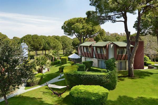 Außenansicht des Gloria Verde Resorts mit Gebäuden, Bäumen und einem weißen Hängematten auf grünem Rasen