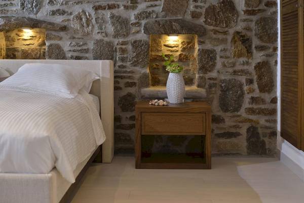 Ein Bett mit weißen Laken neben einer Steinwand und einem Nachttisch mit Vase.