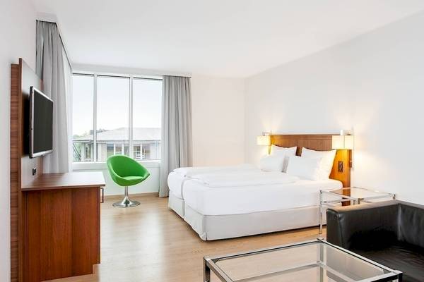 Innenansicht eines Hotelzimmers im NH Hamburg Altona mit zwei Betten, Fernseher, Schreibtisch und grünem Stuhl.