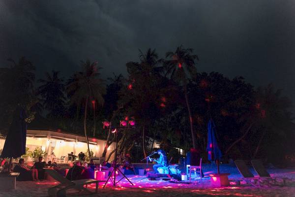 Ein Abendkonzert im Malahini Kuda Bandos Resort mit einem Musiker auf der Bühne und Gästen, die auf Strandliegen sitzen.