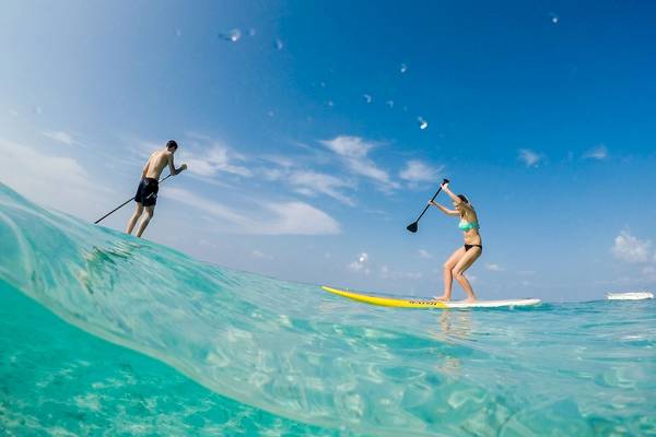Zwei Personen surfen im Meer bei klarem Himmel mit kleinen Wellen und einem Boot im Hintergrund.