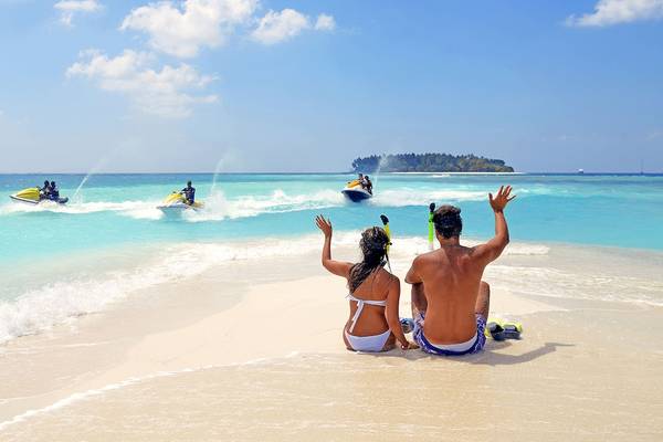 Ein Paar sonnbadet am Strand des Malahini Kuda Bandos Resorts, während Jetskis im Hintergrund fahren.