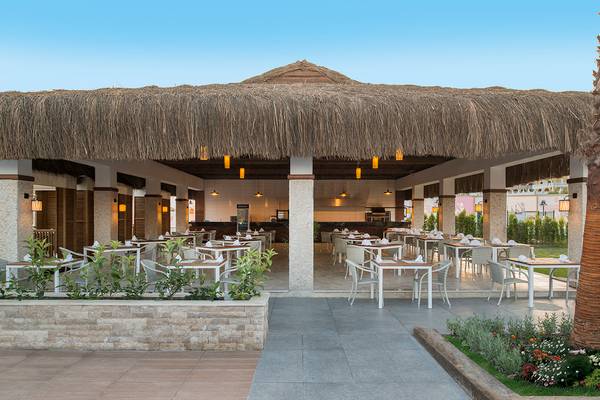 Außenbereich des Aska Lara Resort & Spa mit rustikalem Strohdach und runden Tischen und Stühlen.