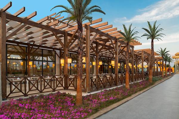 Außenbereich des Aska Lara Resort & Spa mit hölzerner Überdachung, Tischen und Palmen.