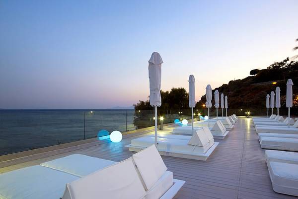 Außenterrasse des Dimitra Beach Hotel & Suites mit weißen Liegen und Sonnenschirmen am Abend mit Blick auf das Meer.