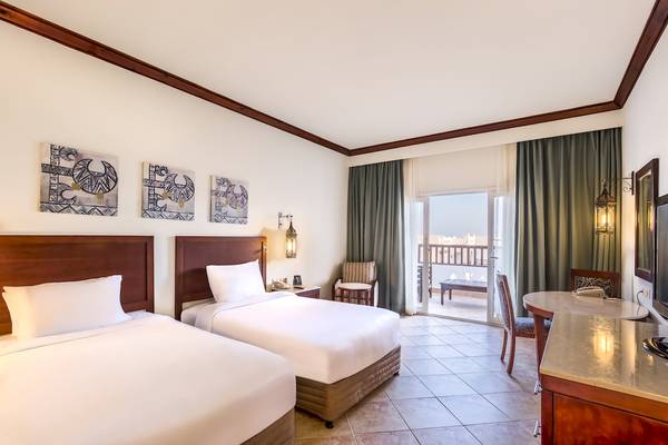 Innenansicht eines Doppelzimmers im Hilton Marsa Alam Nubian Resort mit zwei Betten, einem Tisch, Stühlen und einem Balkon.