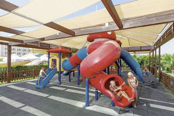 Kinder spielen auf einem Spielplatz mit Rutschen und einem Spielhaus unter einem Sonnensegel.