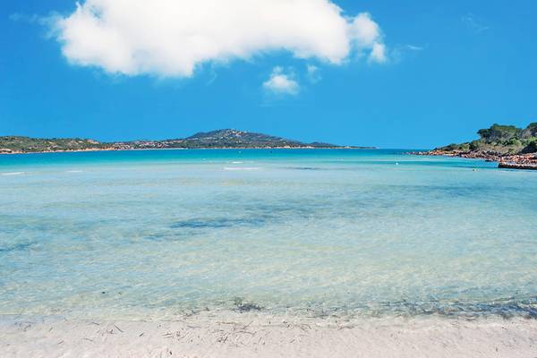Strand am Grande Baia Resort & Spa in Sardinien mit kristallklarem Wasser und Bergen im Hintergrund.