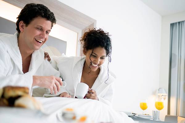 Ein Paar genießt Frühstück im Bett im Novotel London Blackfriars, beide tragen weiße Bademäntel, mit Kaffee, Orangensaft und Frühstücksbuffet im Hintergrund.