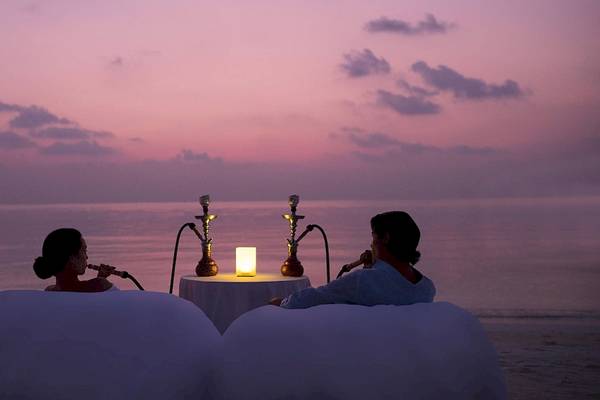Zwei Personen sitzen auf weißen Liegen am Strand des Anantara Dhigu Maldives Resorts und rauchen Shisha bei Sonnenuntergang.