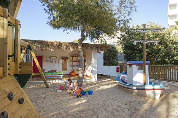Zwei Kinder spielen auf einem Sandspielplatz im Freien, vor einem Baum und einer kleinen Hütte, im Grecian Sands Hotel.