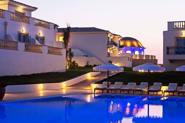 Der Poolbereich des Mitsis Laguna Resorts bei Nacht mit beleuchteten Gebäuden und Liegestühlen.