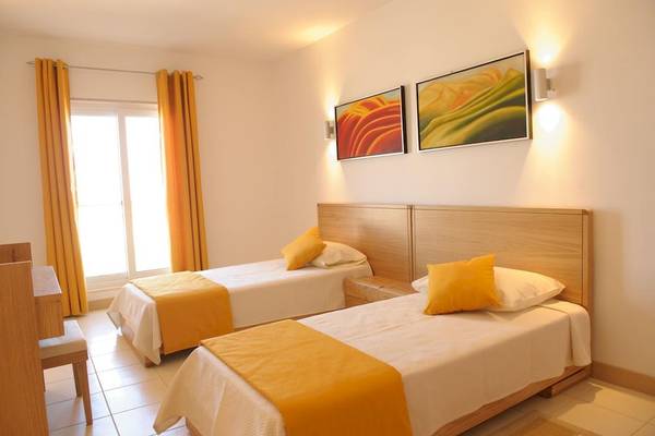 Zimmer mit zwei Betten im Agua Hotels Sal Vila Verde, beide mit gelben Kissen und weißen Laken, neben einem Fenster mit gelben Vorhängen.