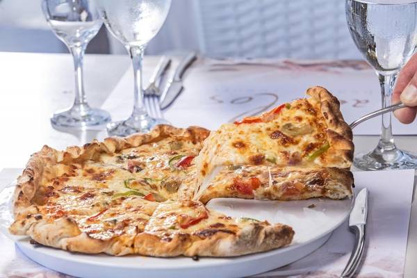 Pizza auf Platte mit Gläsern und Besteck, eine Scheibe wird herausgenommen.