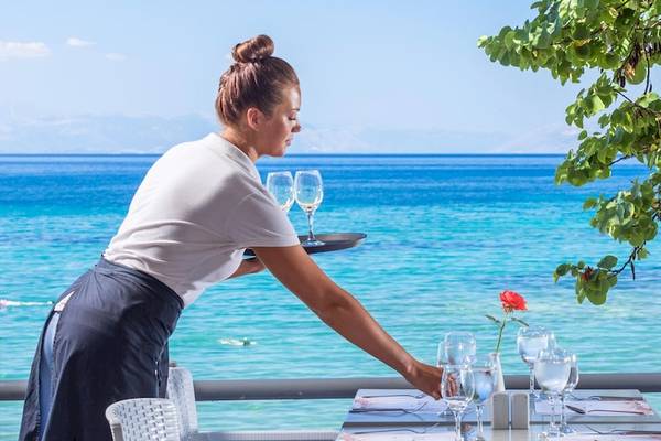 Eine Frau serviert ein Tablett mit Gläsern auf einer Terrasse mit Blick auf das Meer.