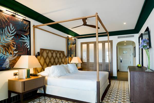 Schlafzimmer im H10 Estepona Palace mit Himmelbett, weißen Laken, zwei Nachttischen und tropischen Wandgemälden.
