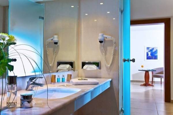 Apollonia Beach Resort & Spa Badezimmer mit zwei Waschbecken, Spiegel, Blumenvase, Handtuchtrocknern und Toilettenartikeln.