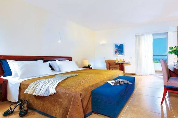 Innenansicht eines Zimmers im Apollonia Beach Resort & Spa mit einem großen Bett, Tisch, Stuhl und Aussicht auf das Meer.