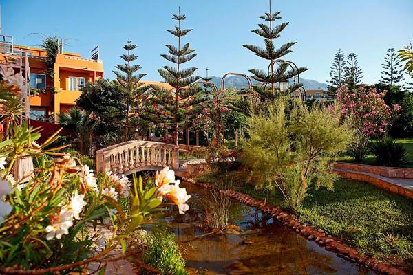 Garten des Orpheas Resorts mit Brücke und Wasser, umgeben von Pflanzen und Bäumen.