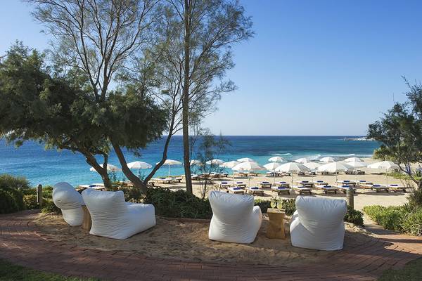 Außenbereich des Grecian Sands Hotels mit weißen Liegestühlen und Sonnenschirmen am Strand.